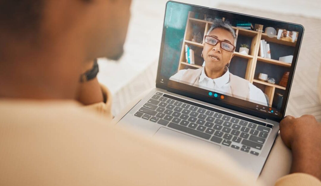 A man sits with a laptop on his legs. A video call with a health-care professional sitting in her office is on the screen.