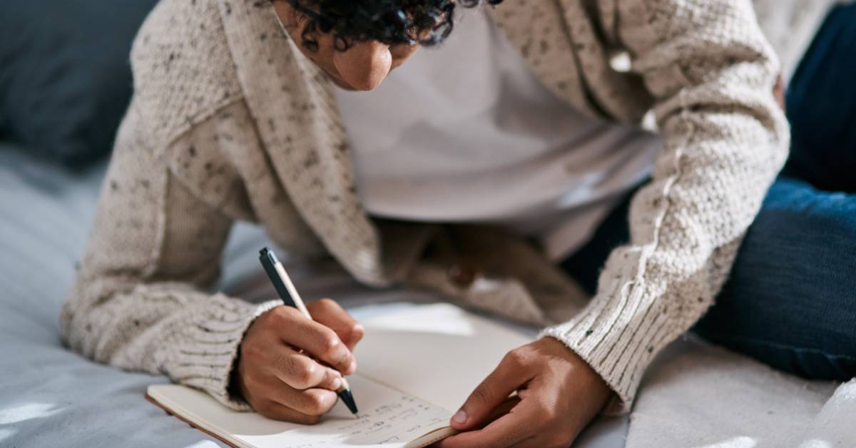 A young man wearing a beige cardigan and white tee rests his elbow on the bed and writes notes in a lined journal.