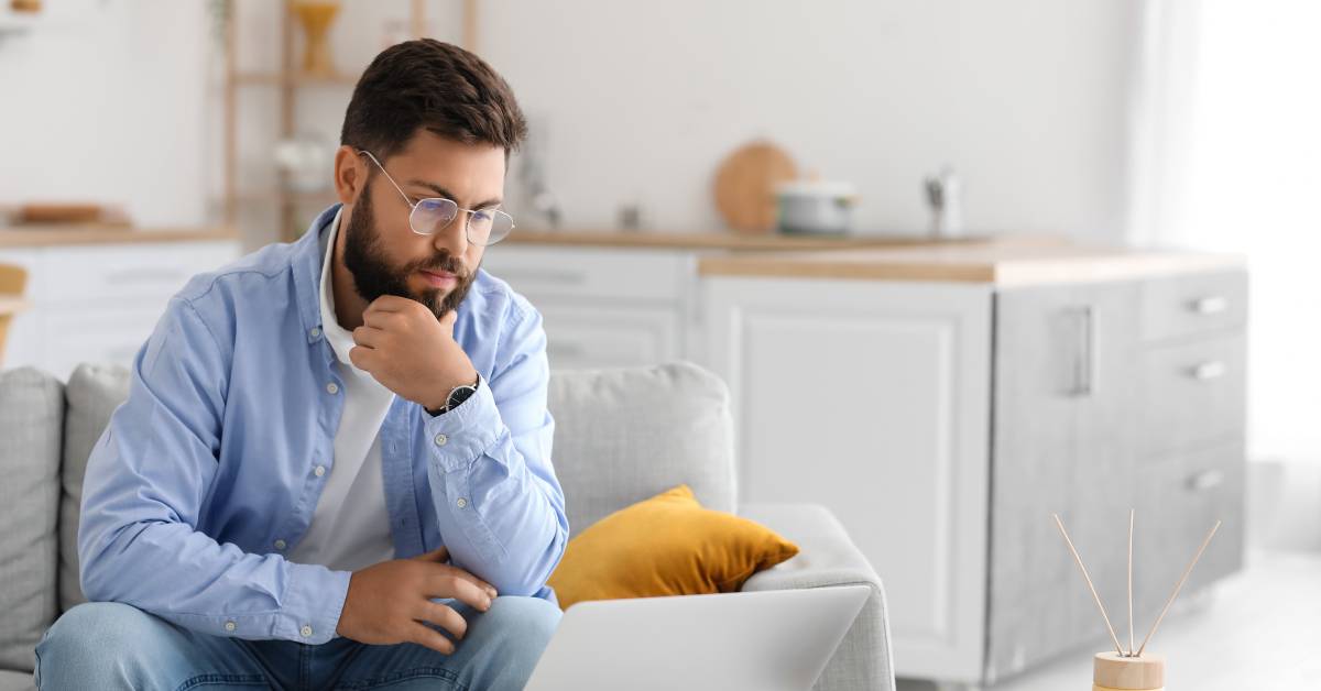 A man sits on his couch at home with a computer, notebook, and pen on a white coffee table. He looks down at the screen.