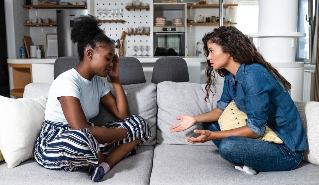 Two female friends sit cross-legged on a gray couch facing each other while having a serious conversation.