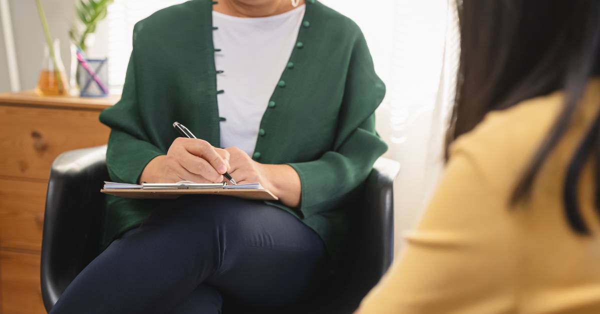 A teenage girl wearing a yellow shirt faces a therapist sitting in a black chair with a clipboard wearing a green cardigan.