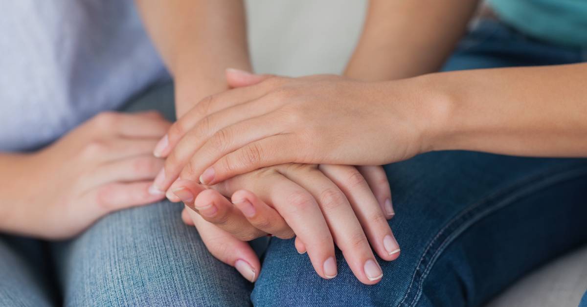 Two female friends sit close together on a couch, one embracing the other's hand for comfort.