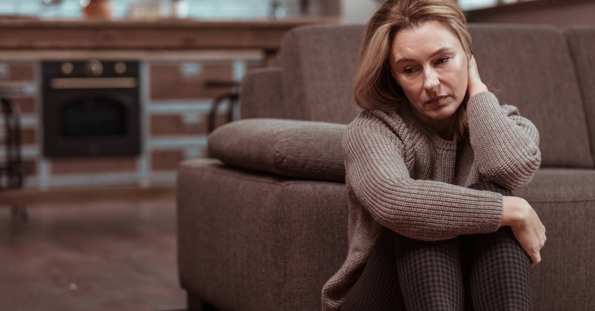 A woman wearing a brown sweater sits on her living room floor near her couch with her knees to her chest looking depressed.