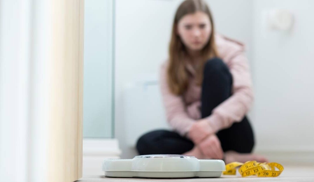 A teenage girl sitting on her bathroom floor staring at a scale and yellow measuring tape in focus in the foreground.