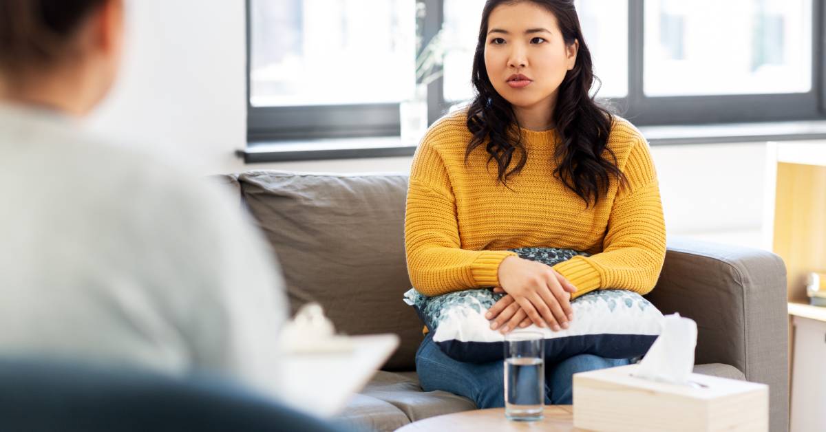 A young woman wearing a yellow sweater sits on a couch across from an out-of-focus psychiatrist in her office.