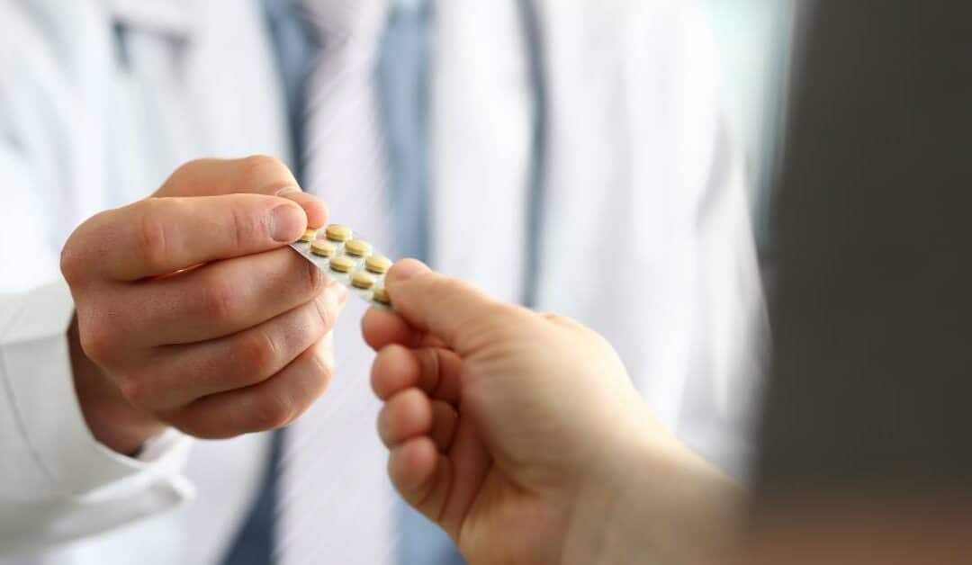 A male doctor wearing a white jacket and tie hands a male patient a blister pack of yellow pills.