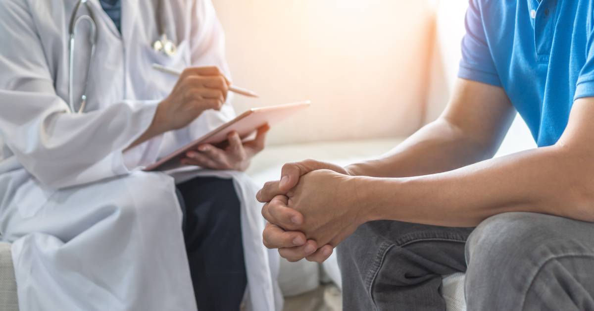 A doctor wearing a white coat sitting and taking notes across from a man wearing a polo shirt and jeans with clasped hands.