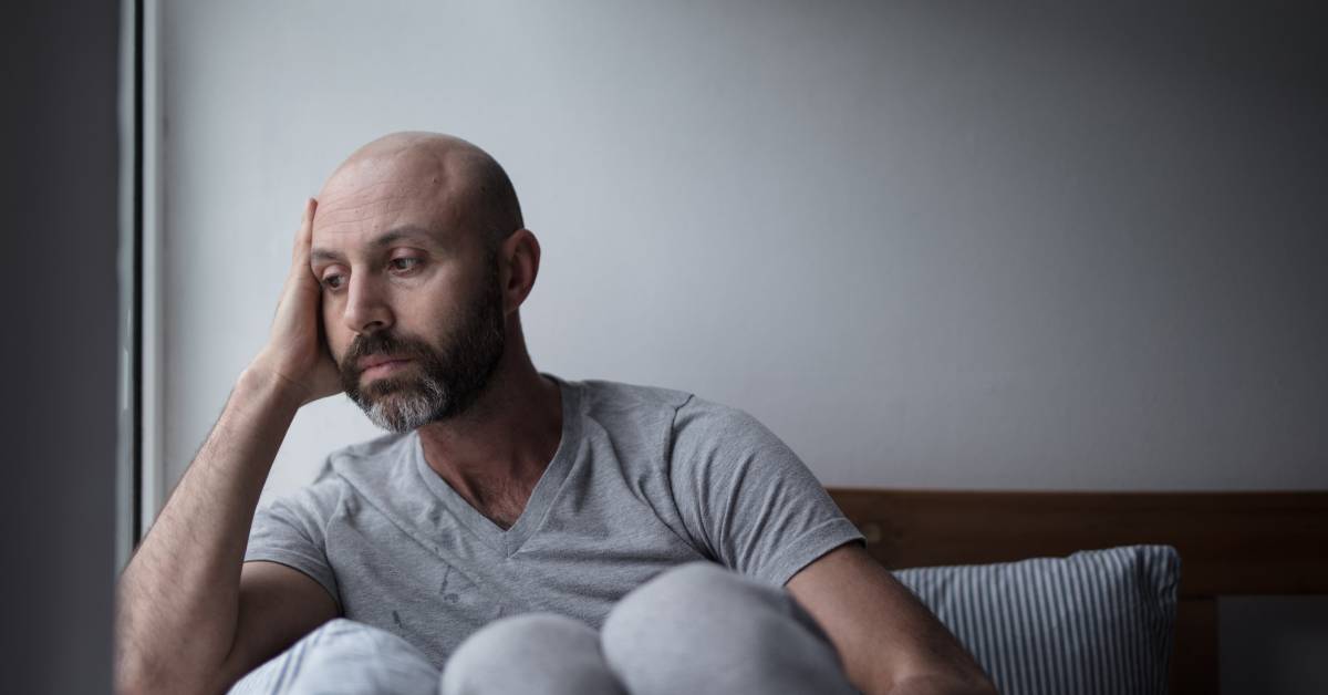 A bald man sitting up in bed with his knees to his chest, resting his elbow on a window ledge, and looking out of his window.