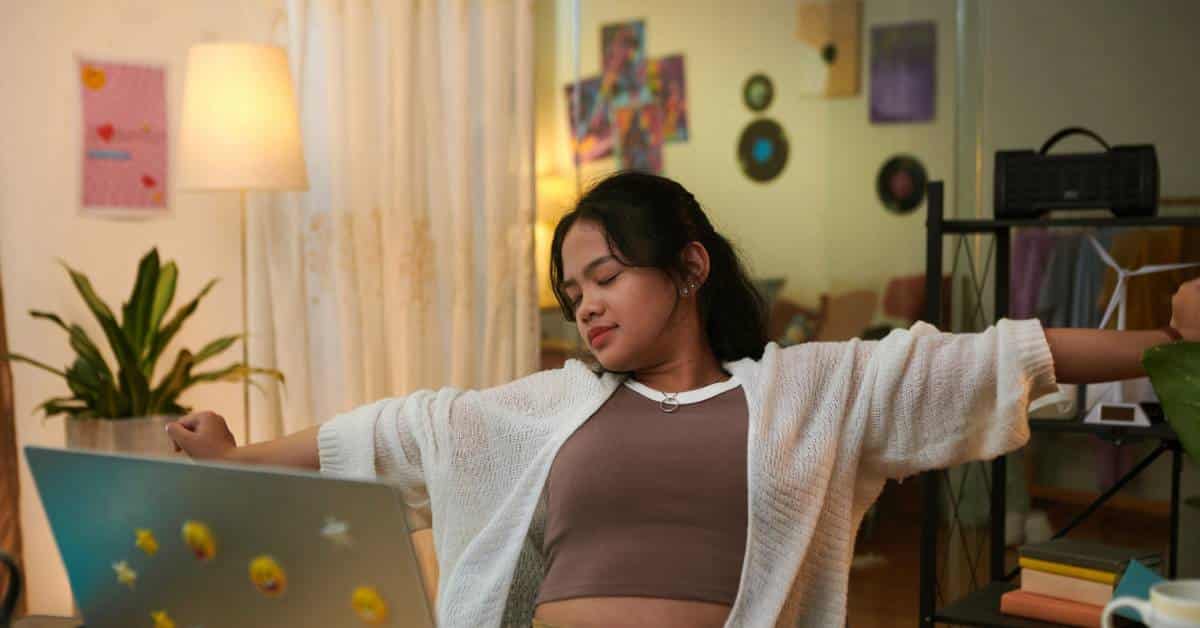 A teenage girl stretches her arms while sitting at her desk in her bedroom, taking a break from school work.