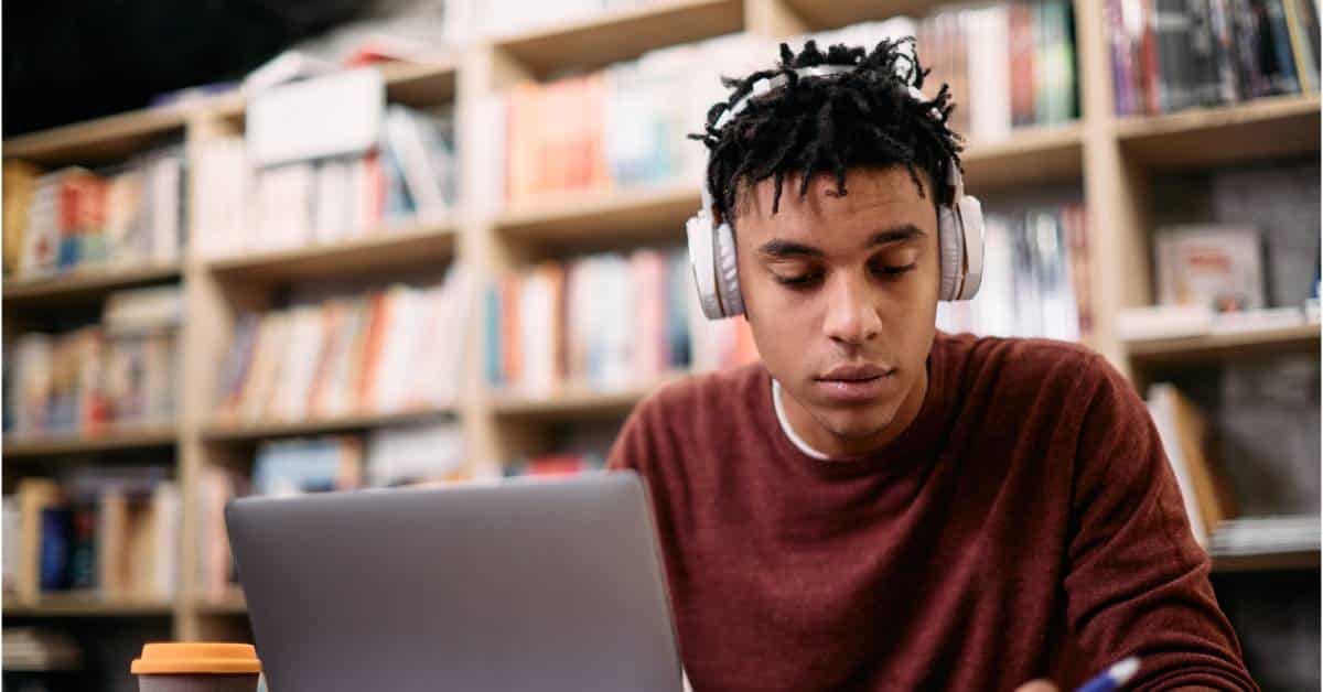A male teenage student wearing headphones studies in a library at a table with at laptop and open notebook.