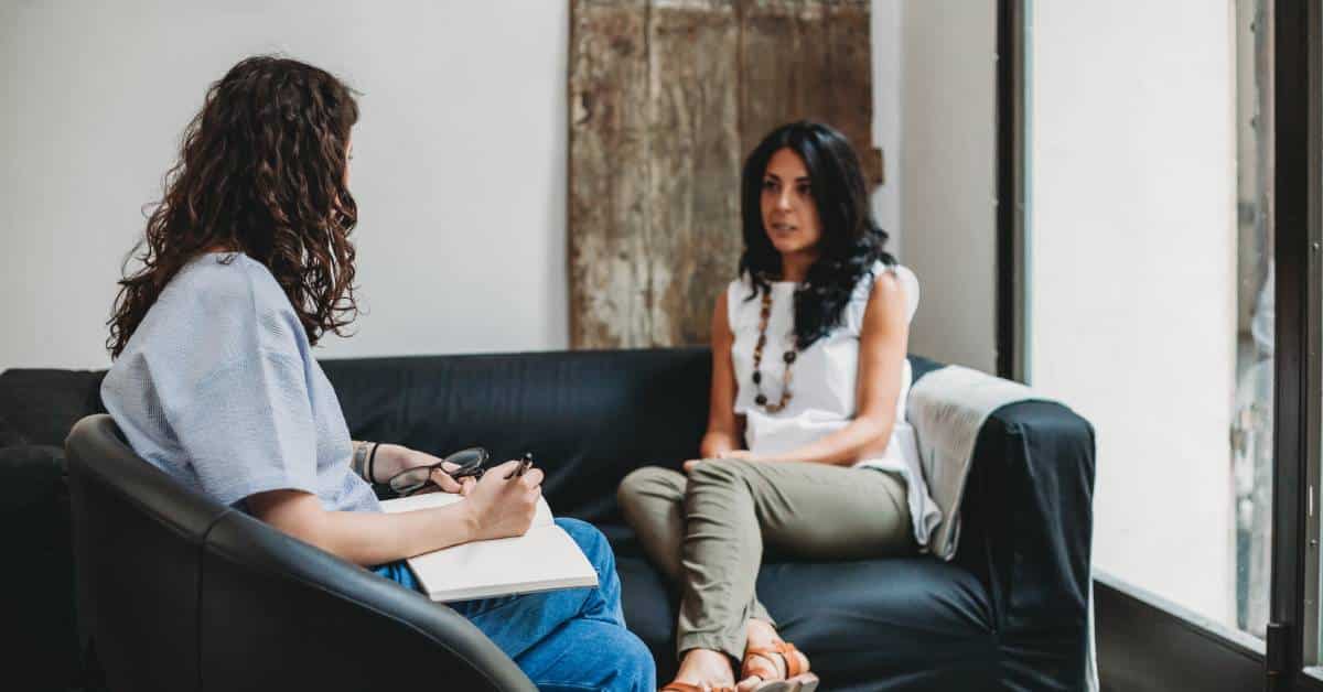 A woman sitting on a navy couch faces a female therapist sitting in a black chair, taking notes in a notebook.