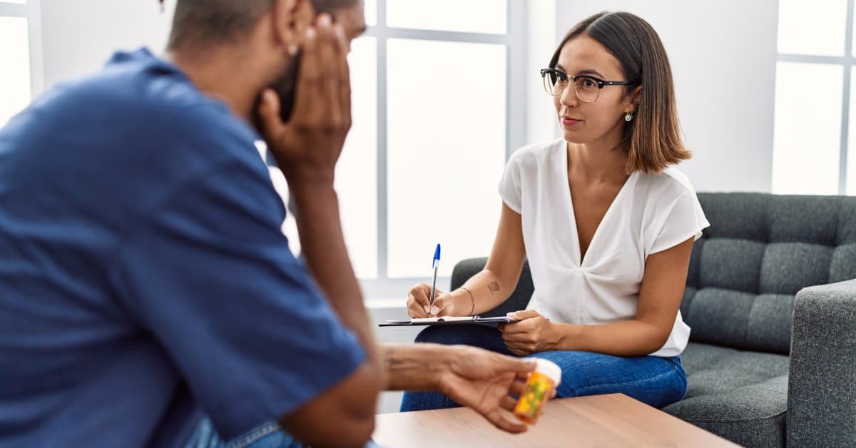 A man holding an orange pill bottle sits across from a female psychiatrist taking notes on a clipboard.