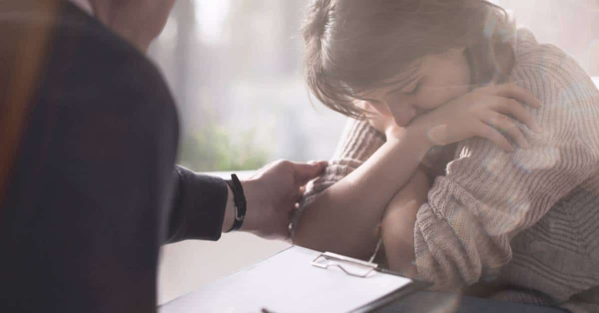 A psychiatrist sits across from an upset woman crossing her arms. The psychiatrist touches her arm for comfort.