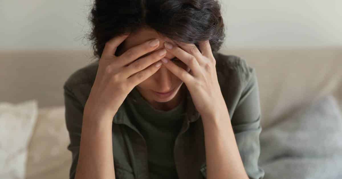A woman with dark hair wearing grey clothing sits on her couch holding her head feeling distressed.