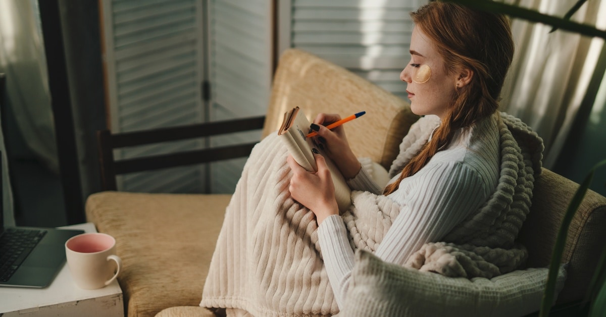 A woman wrapped in a blanket, wearing undereye patches, is sitting on a loveseat and writing in her journal.