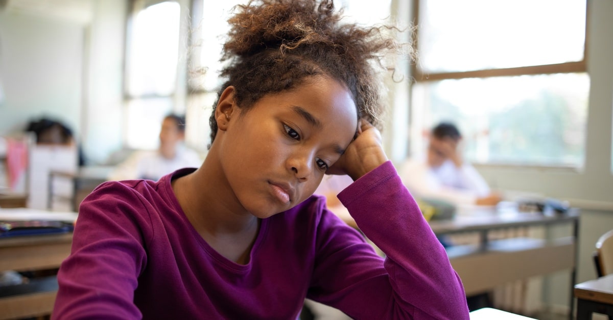 A young girl sitting in a classroom with a sad expression on her face. She rests her head in her hand.