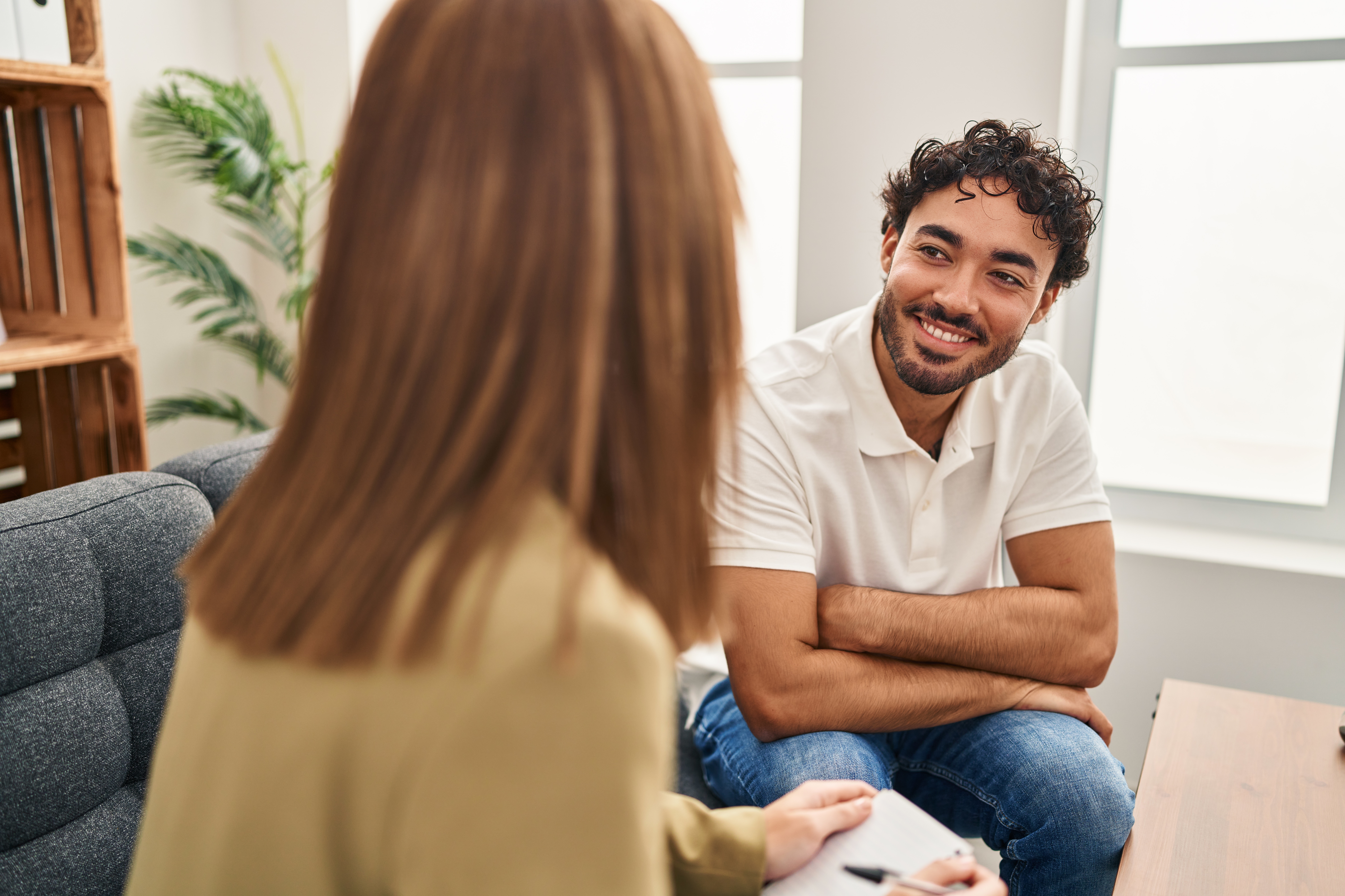 A smiling man and woman psychiatrist holding a clipboard sit facing each other, both sitting on a grey couch.