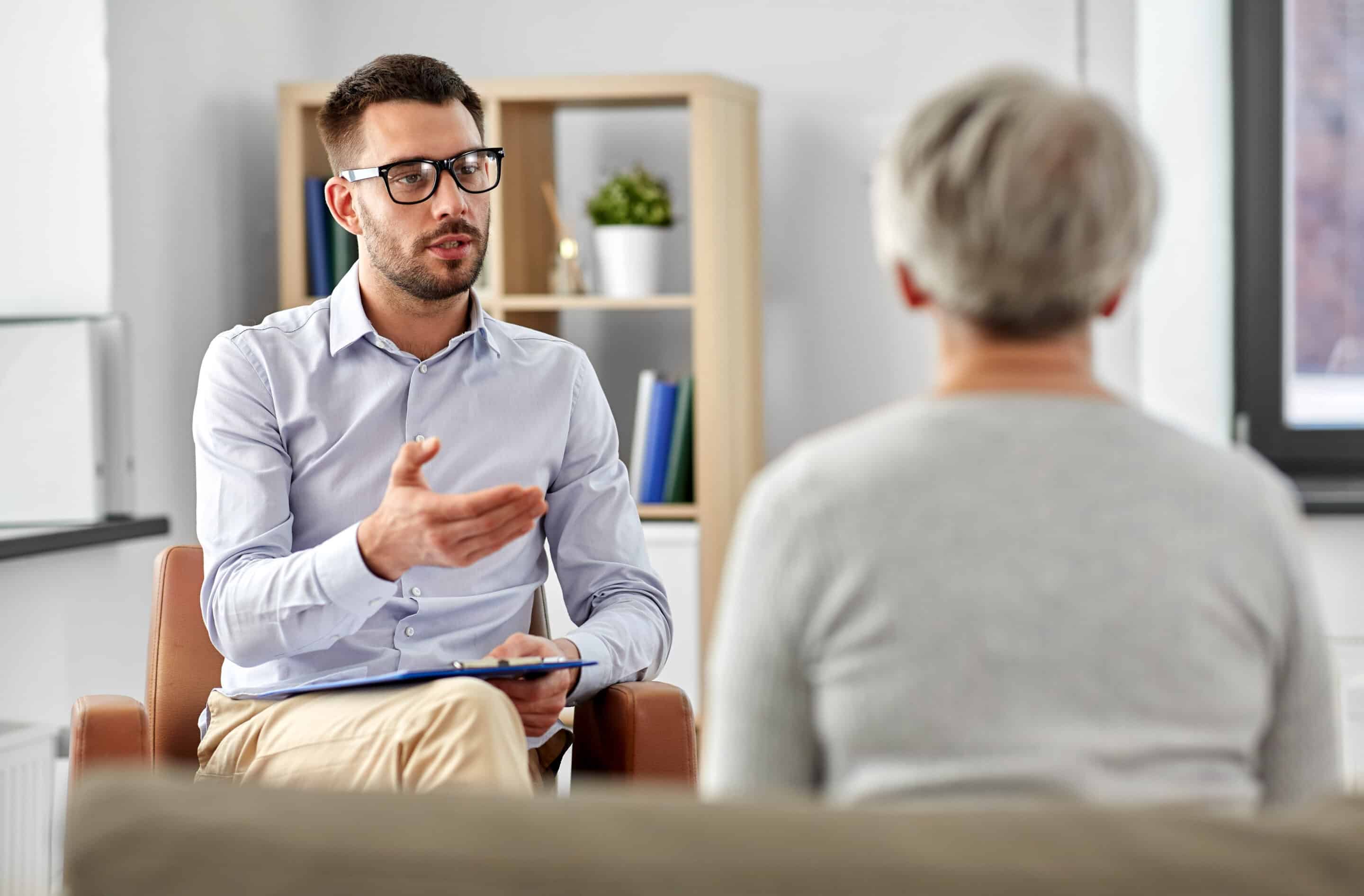 A male psychiatrist holding a clipboard sits across from a senior woman on a couch in the foreground in his office.