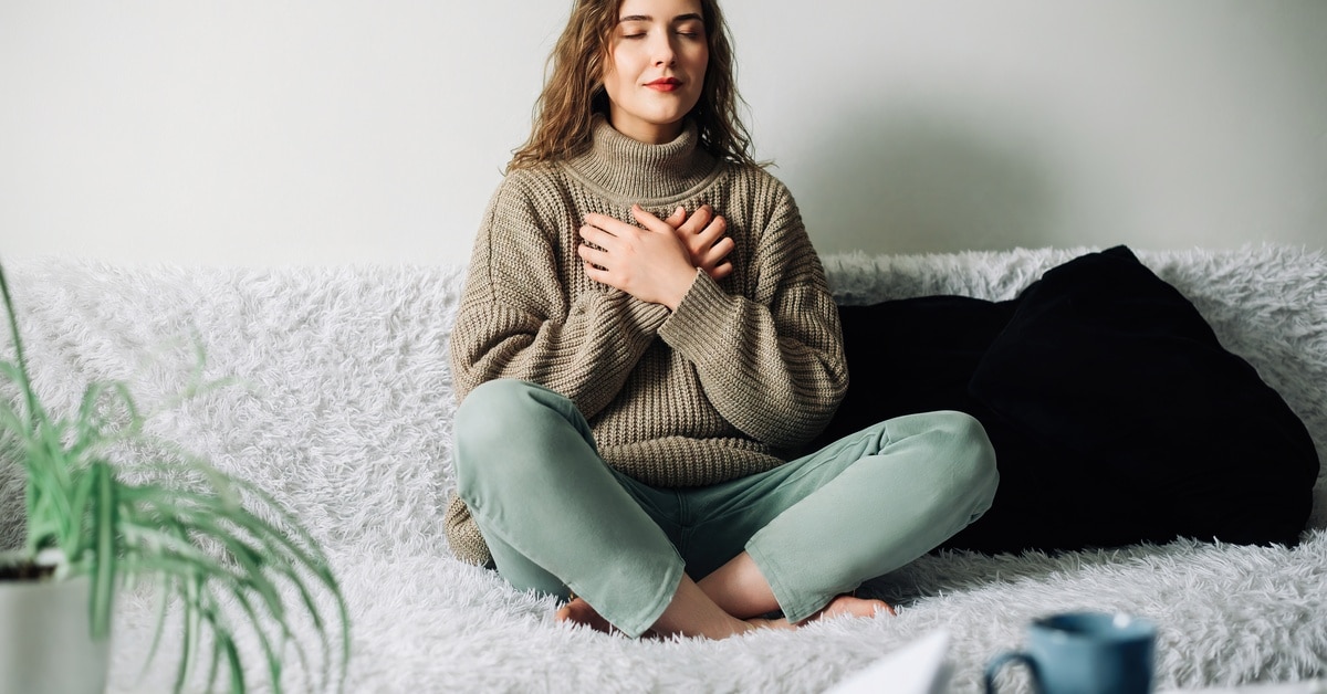 A woman wearing a sweater and jeans sits cross-legged on her fuzzy couch, holding her hands to her chest to remain calm.