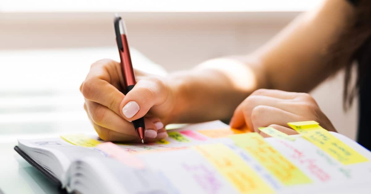 A close-up of a woman's hand writing in a planner with a red and black pen. The planner is covered in multi-colored sticky notes.