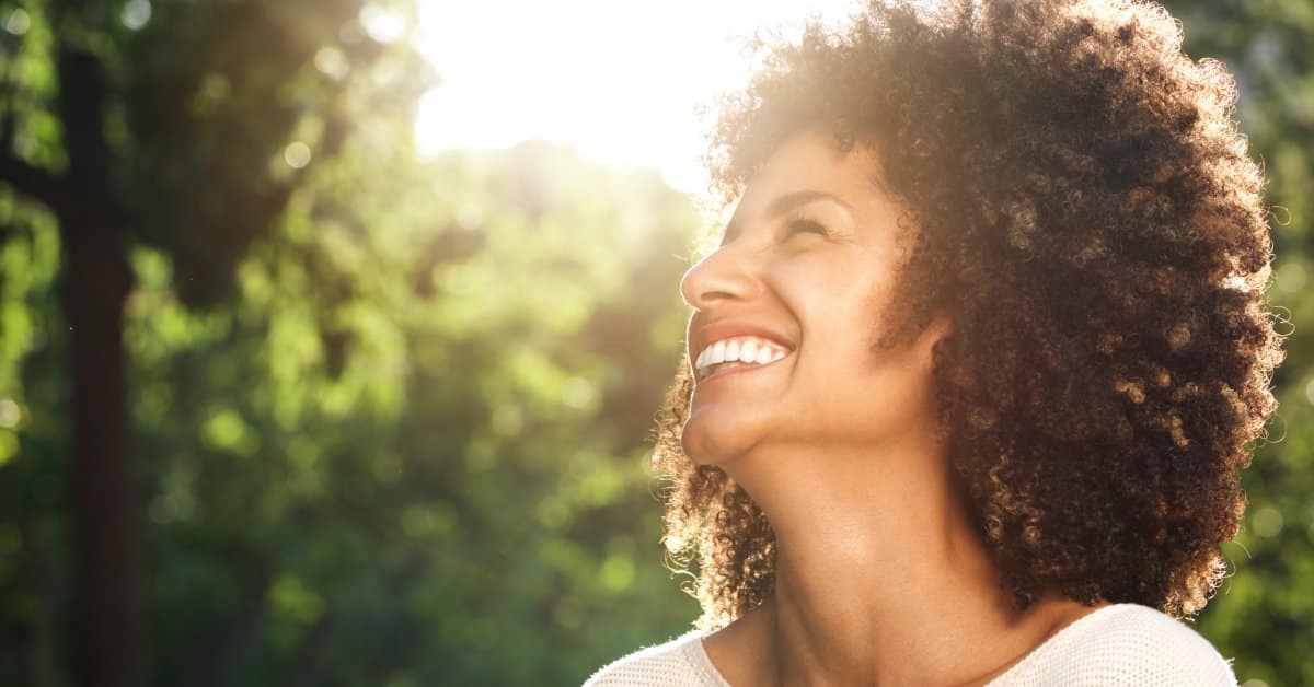 A woman stands outdoors among trees, smiling as she looks up at the sky, with sunlight streaming through the leaves.