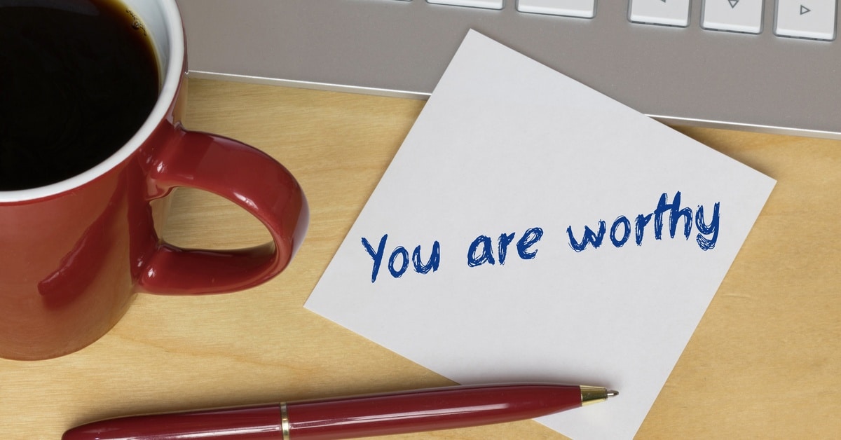 A close-up of a sticky note reading "you are worthy" next to a red pen, red mug with coffee, and a computer keyboard.