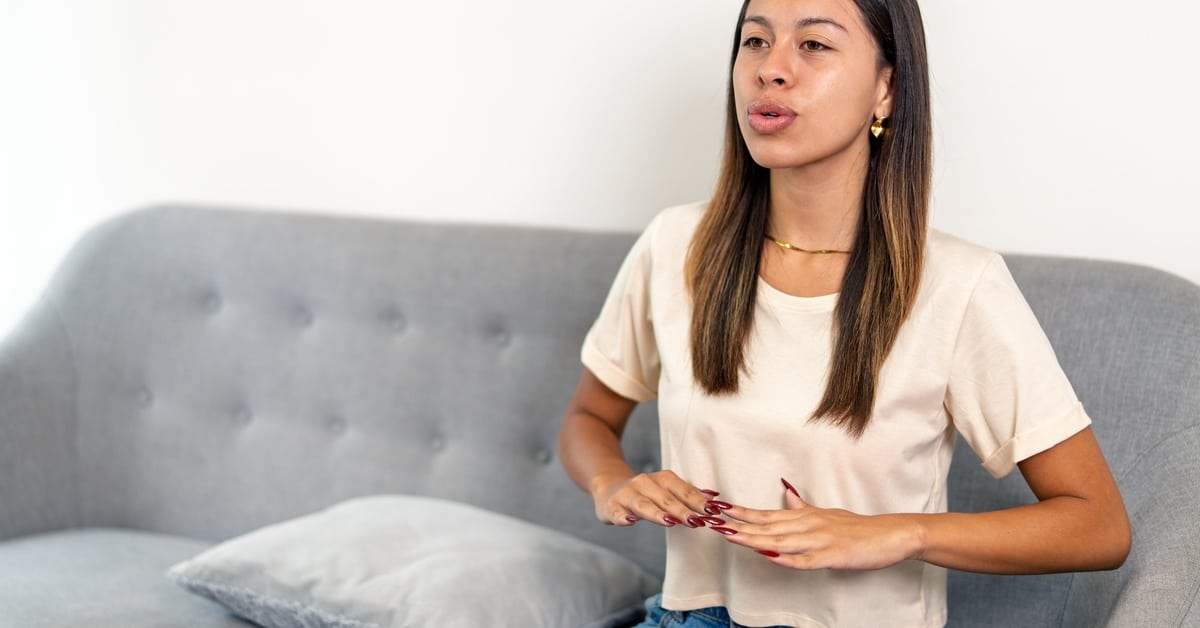 A woman sits on her couch, performing a breathing exercise, while moving her hands downward in front of her chest.