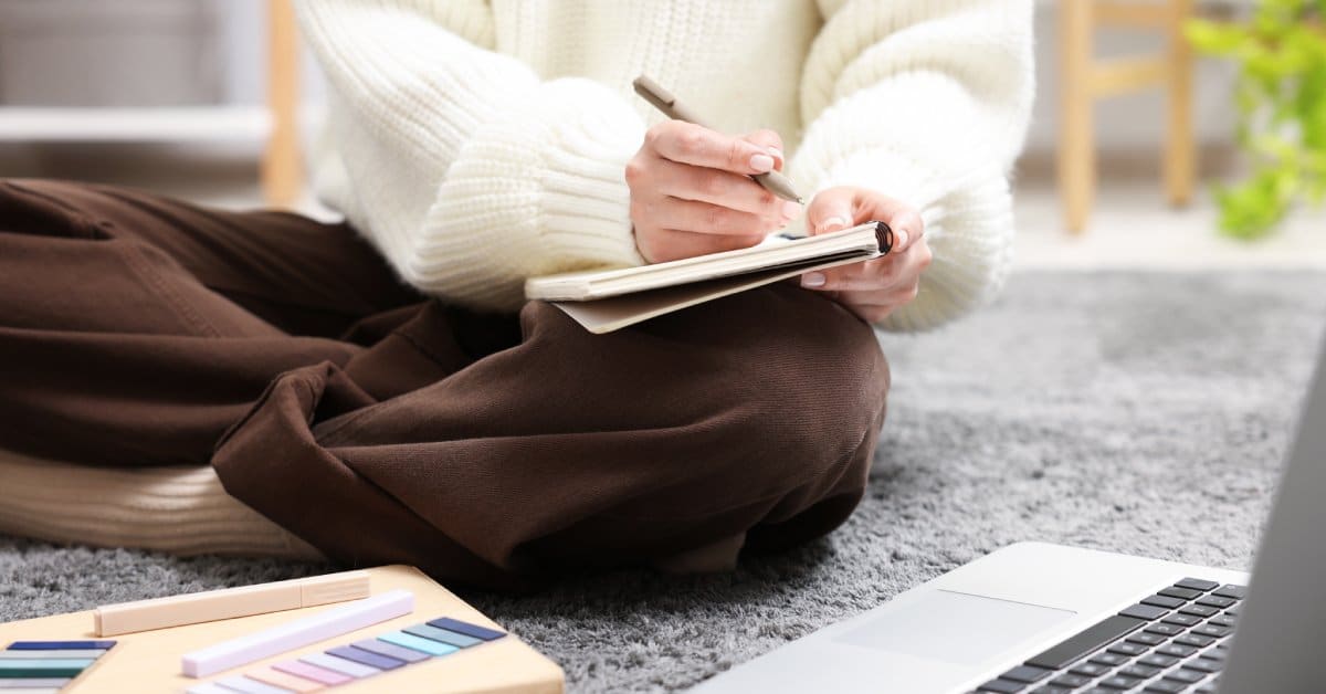 A woman sits cross-legged on the floor, writing in a notebook while facing her laptop, with highlighters and pens beside it.