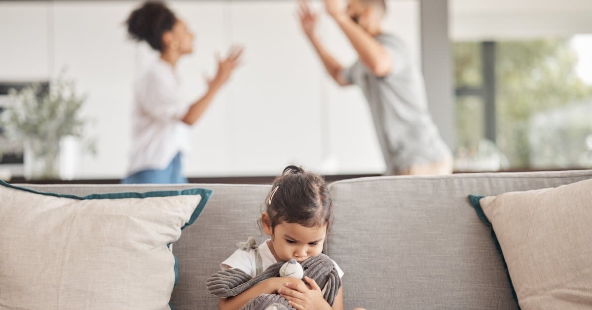An upset young girl sits on a couch holding a stuffed rabbit as her parents fight in the blurred background.