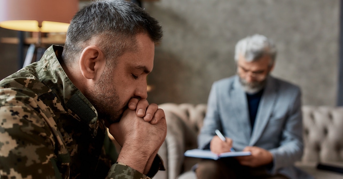 A male soldier wearing camouflage puts his hands to his mouth, while a male psychologist writes in a notebook, sitting on a couch.