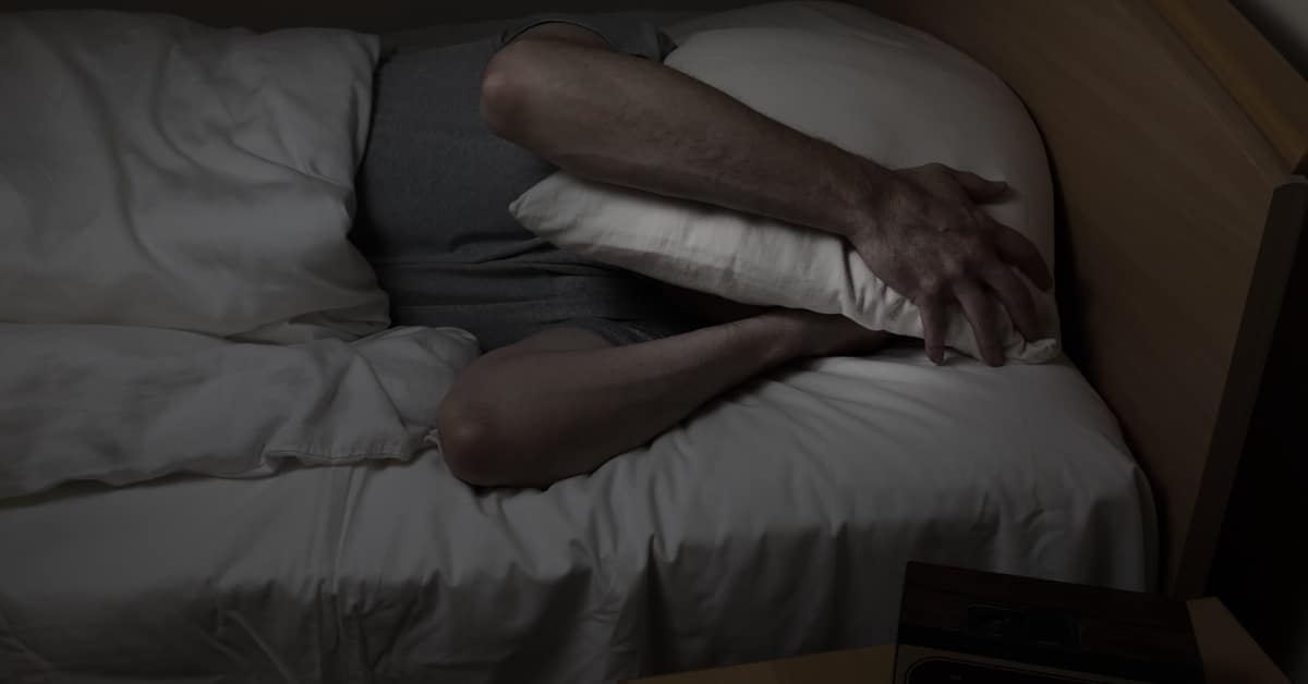 A man wearing a t-shirt lies in bed under a comforter, holding a white pillow over his head, unable to sleep.