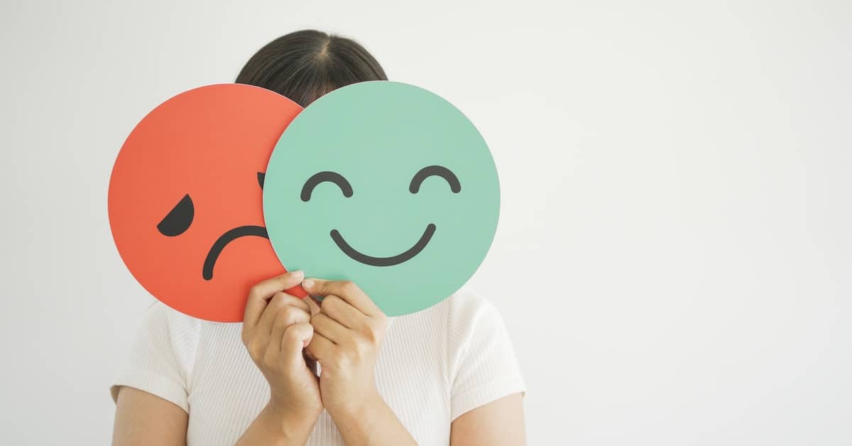 A woman holds two images of faces above her face. One is a red frowning face and the other is a green smiling face.