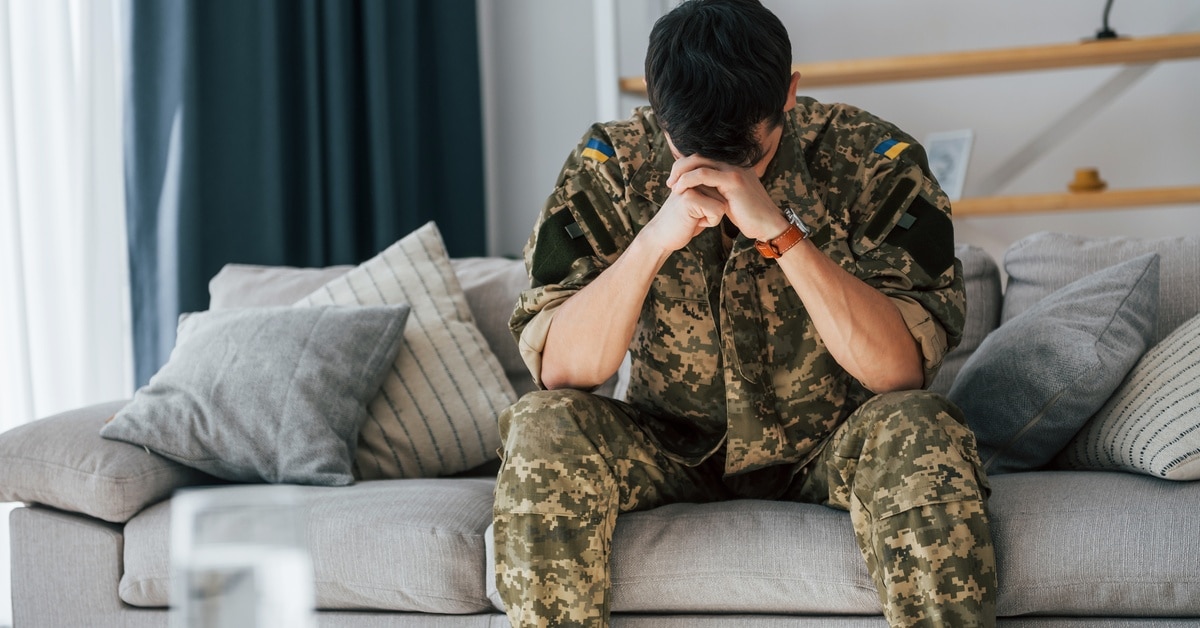 A man wearing a camouflage soldier uniform sits on a couch, resting his head on his clasped hands, appearing anxious.