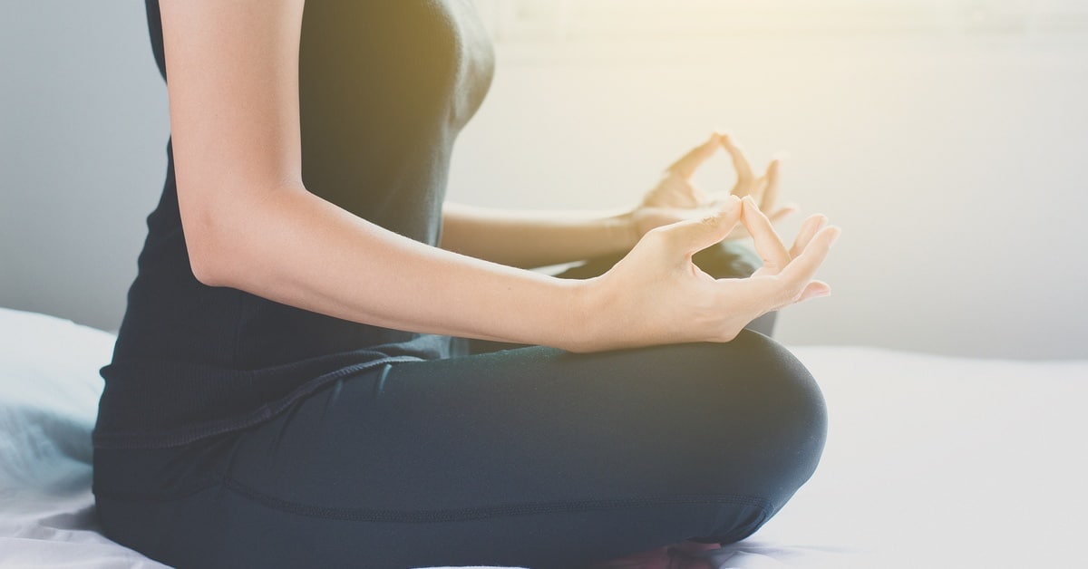 A woman sits cross-legged on a bed with a white blanket. Her hands rest on her knees as she meditates.