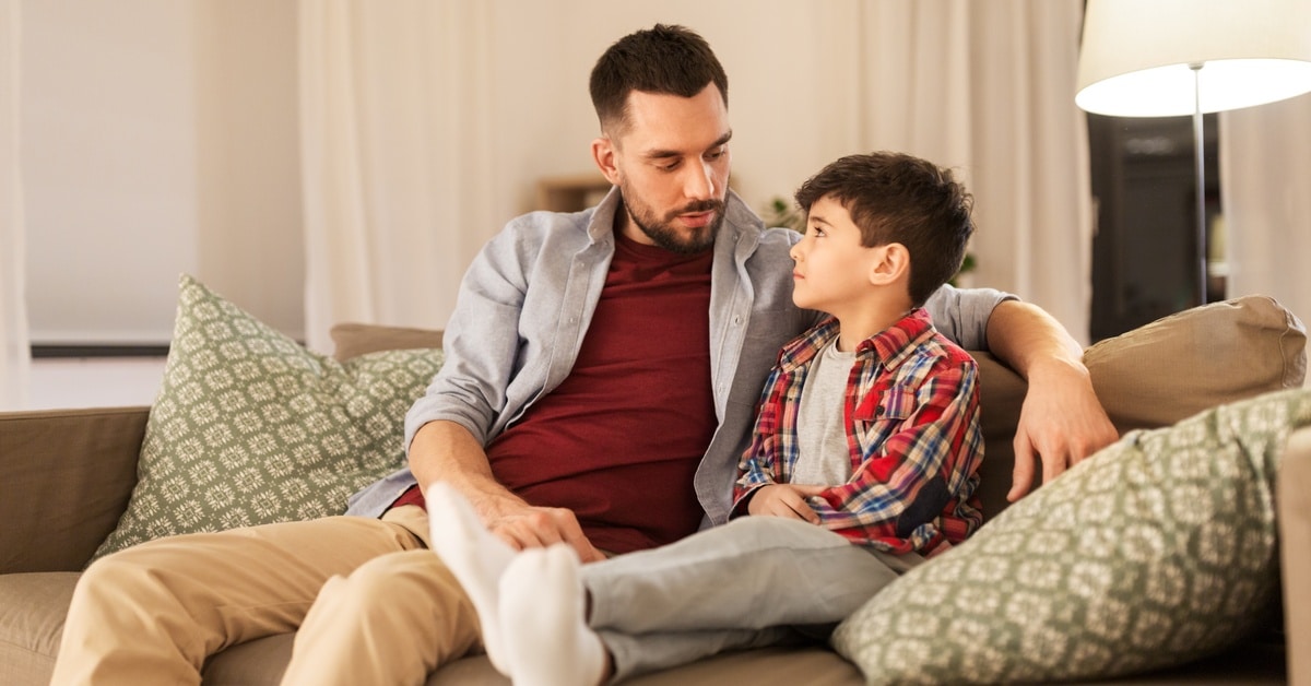 A father and son sitting together on a couch, talking and looking at each other. The father’s arm is around his son.