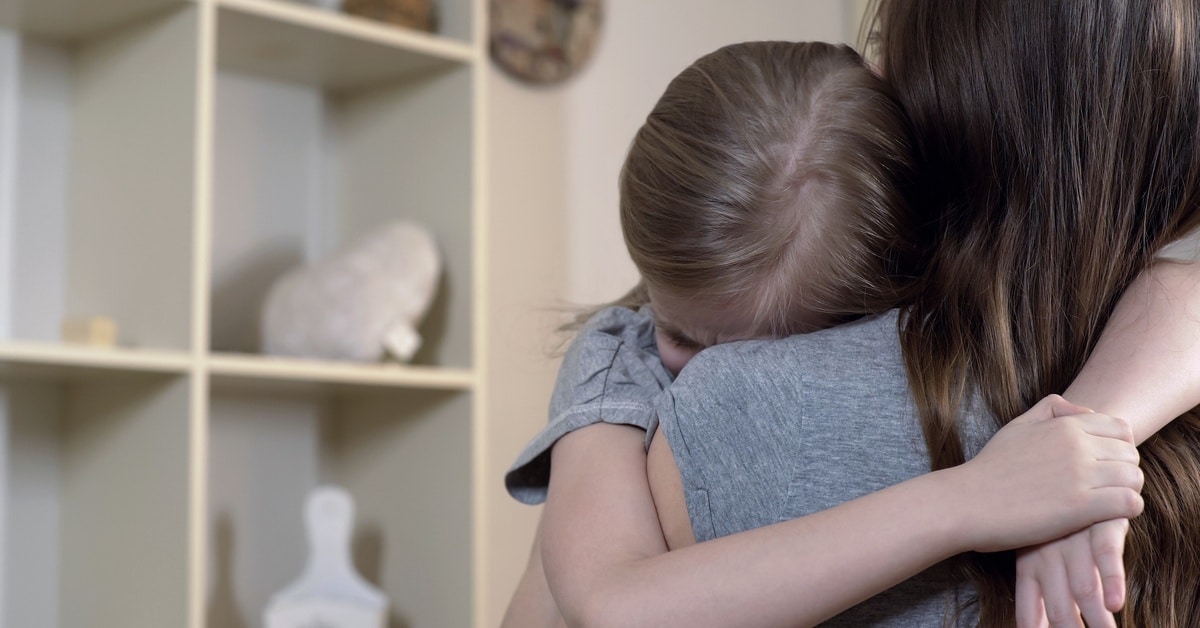 An upset crying girl with her arms wrapped around her mother. The girl rests her forehead on her mom's shoulder.