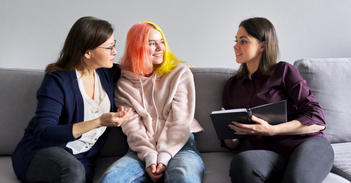 A mother and teenage daughter with dyed hair sit close together on a couch facing a professional woman holding a notebook.