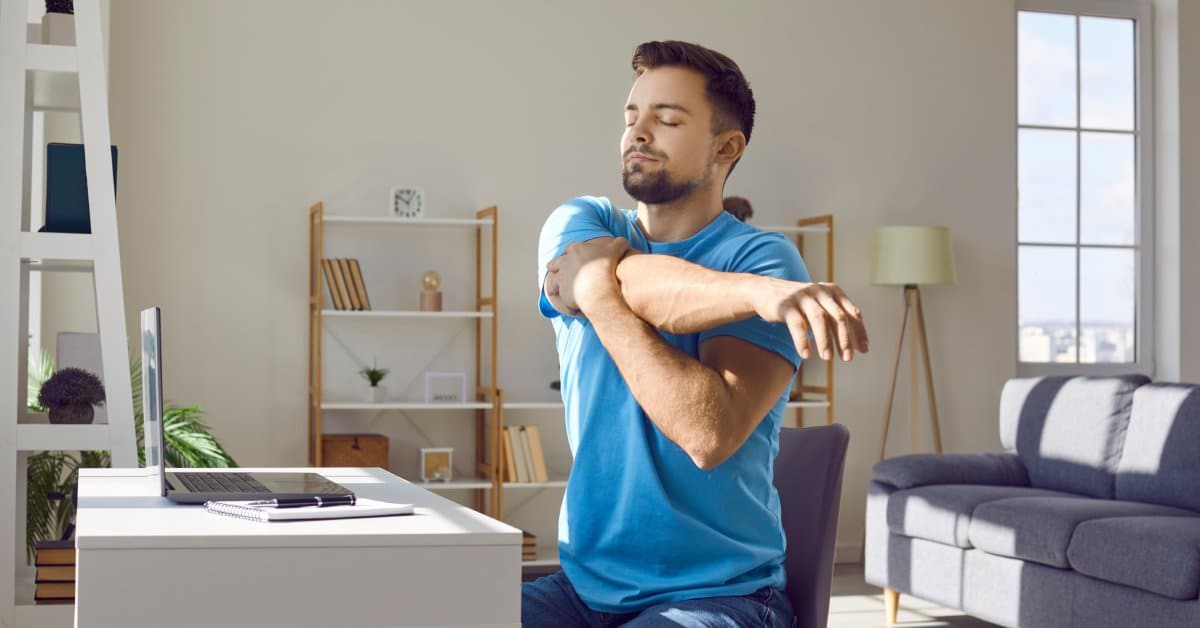 A man is sitting at his desk with a laptop. He stretches his right arm across his body with his eyes closed.
