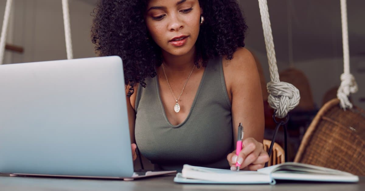 A young woman sits at a table with her laptop and a planner. She writes in her planner with a pink pen in her left hand.