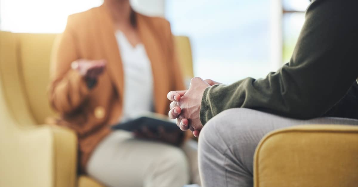 Two people sit across from each other in yellow chairs. One person clasps their hands, the other speaks and holds a book.