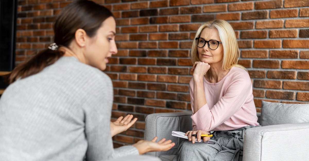 Two women sit across from each other in cushioned chairs. One woman holds a notebook and listens as the other speaks.