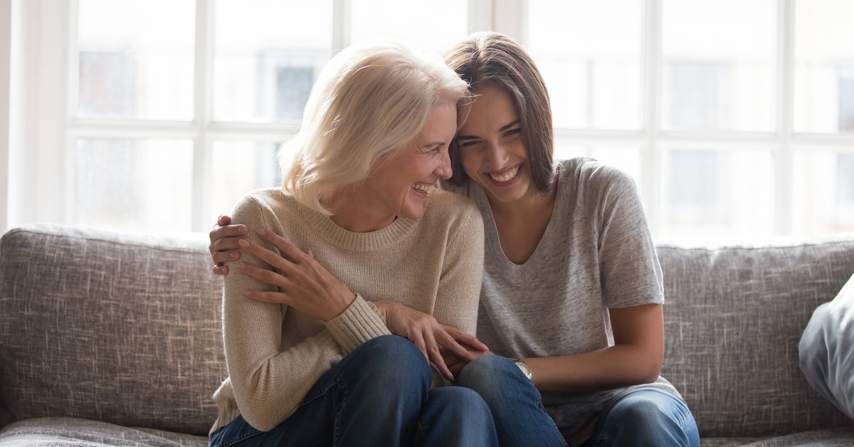 Two women are laughing and sitting on a gray couch. The younger woman wraps an arm around the elder woman.