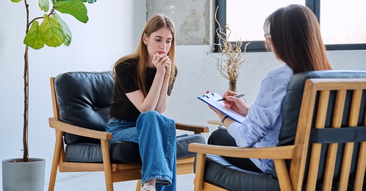 Two women sit in wooden chairs with black cushions in a bright office. One woman holds a clipboard and a pen.