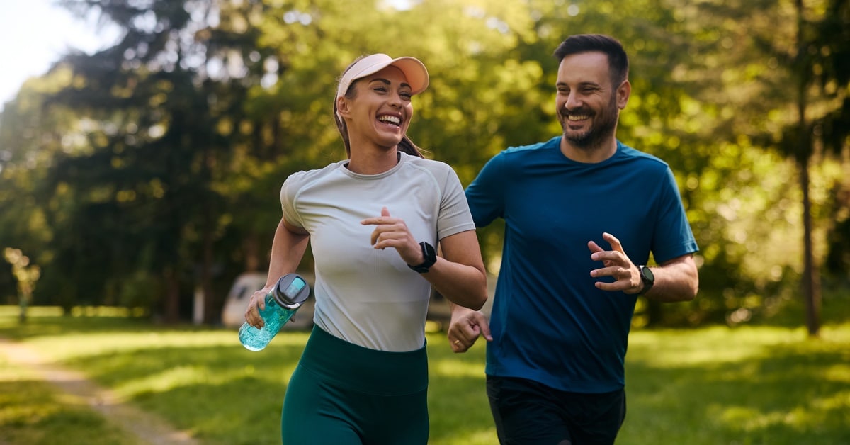 A woman and a man are smiling and jogging outside. A ray of sunlight passes through the tall green trees.