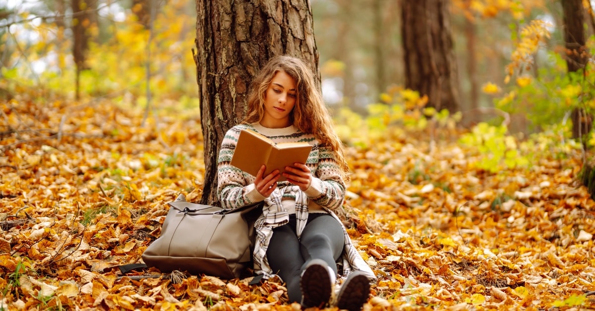 A woman with curly hair sits against the trunk of a tree and reads a book. Orange and yellow leaves cover the ground.