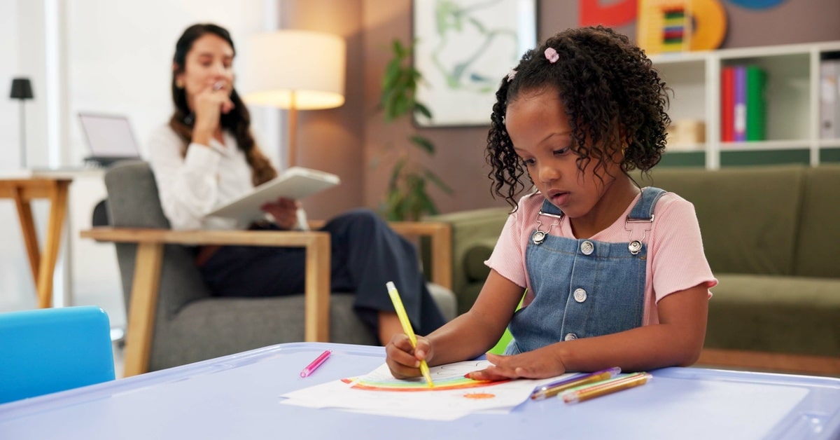 A young girl at a table colors a rainbow with a yellow marker. A young female therapist sits in the background.