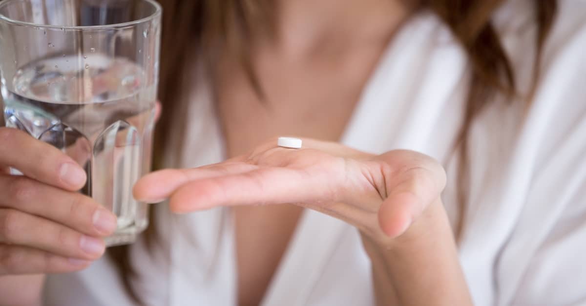 A close-up of a woman's hands. One hand palm side up holds a small, white pill, the other holding a glass of water.