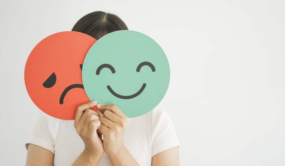 A person holds two signs in front of their face. One is red with a frowny face, and the other is green with a smiley face.