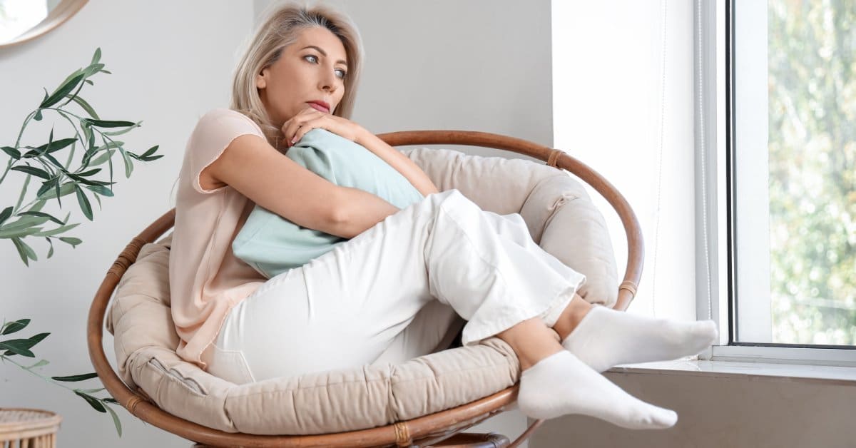 A woman holds a pillow and sits sideways in a round, wooden chair with a white cushion. She looks out the window.