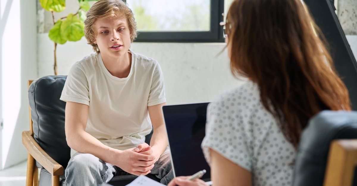 Two people sit across from each other in an office. One adult is writing in a notebook and listening to the other individual.